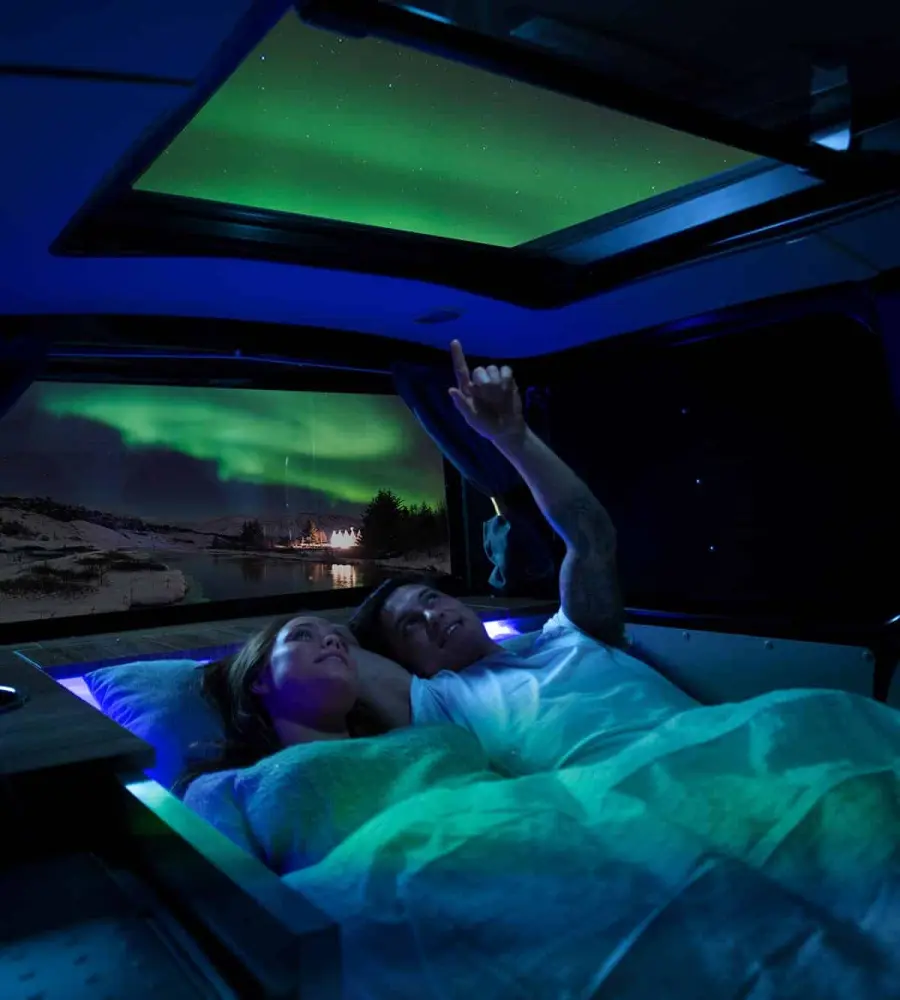 Two travellers in bed in a Cozy camper, pointing at the northern lights through the open sky roof