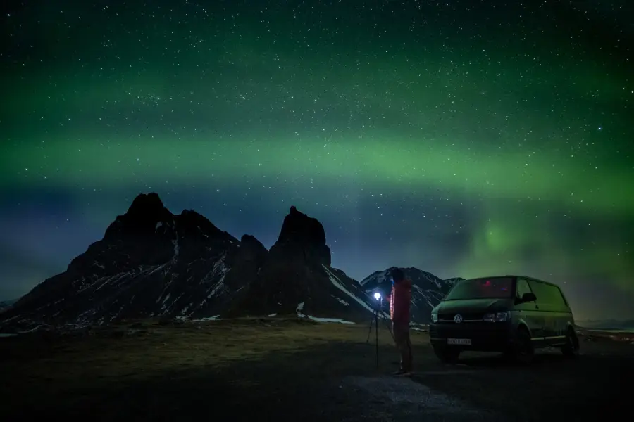 A photographer with a tripod beside a camper, under the northern lights at Vestrahorn