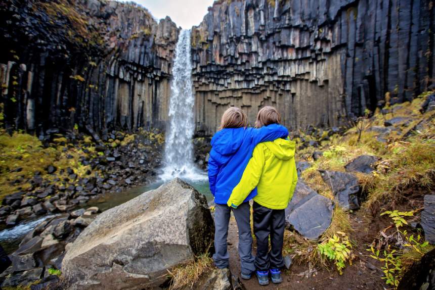 Two kids stand together, looking at a high waterfall in Iceland.