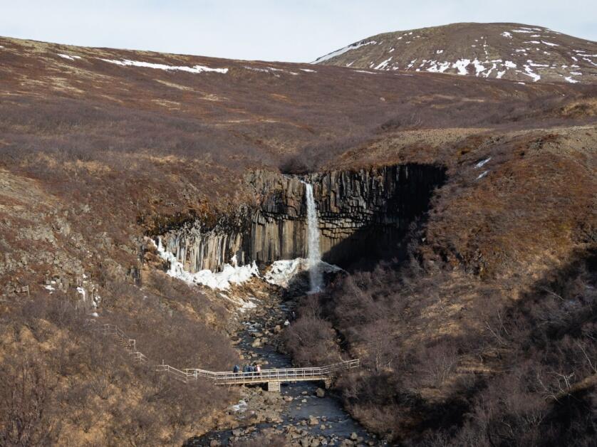 A distant shot of Svartifoss, a waterfall in Skaftafell’s National Park, its basalt columns barely visible and surrounded by golden grassy hills.