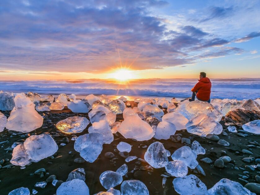 A person sets on a massive ice-chunk on Diamond Beach, strewn with more ice. The sun is golden yellow, shining from under feathery clouds and the color is reflected in the ice.