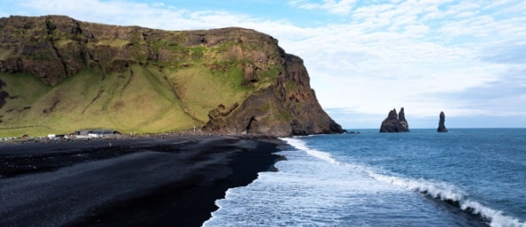 An aerial view of Reynisfjara black sand beach, with waves foaming white along the dark shore and grassy hills. Basalt rocks jut from the ocean.