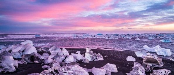 Diamond Beach, Iceland, at sunset. The sky is streaked with purples, pinks and blues while icebergs resting on pitch black sand reflect the sky’s colors.