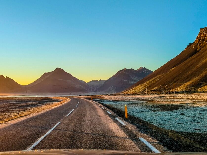 Golden sunlight streams from the left-hand side and spills onto a curving highway with Iceland’s hills and mountains in the distance.