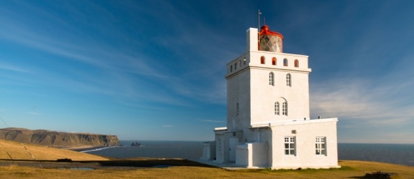 Iceland’s Grotta lighthouse is standing against black sand beaches surrounded by wild grass. The ocean and rolling hills are in the distance.