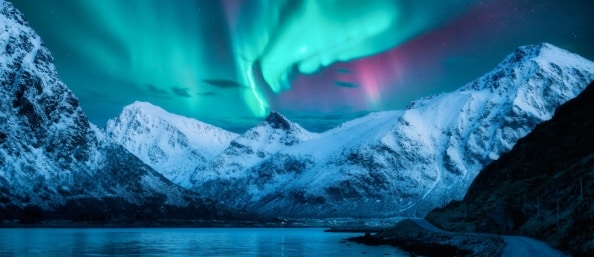 The Northern Lights, a curtain of green, blue, and purple, hangs over a snowy mountain, cradling water that reflects the sky.